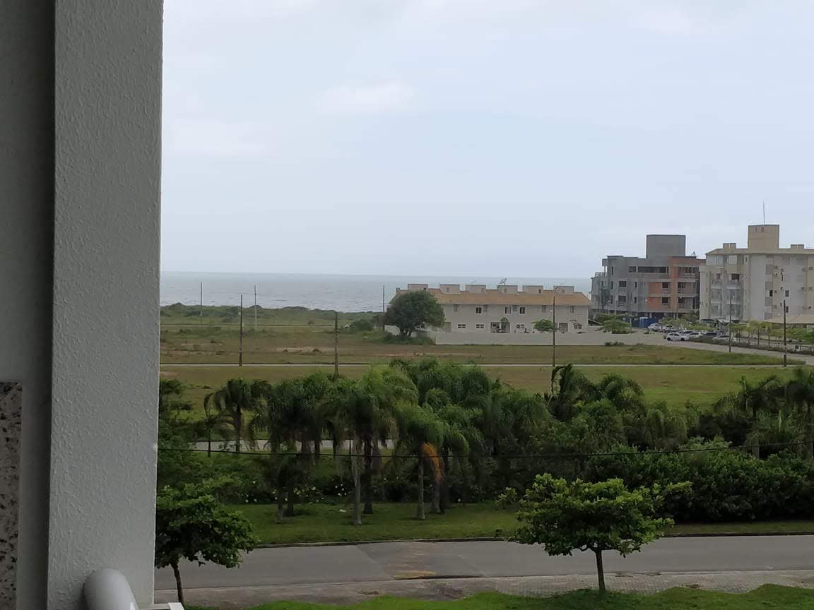 A coastal view is presented from a third-floor balcony, showcasing the sea in the distance along with a mix of residential buildings and green landscaping. Palm trees and neatly trimmed grass add to the scene, creating a serene atmosphere.