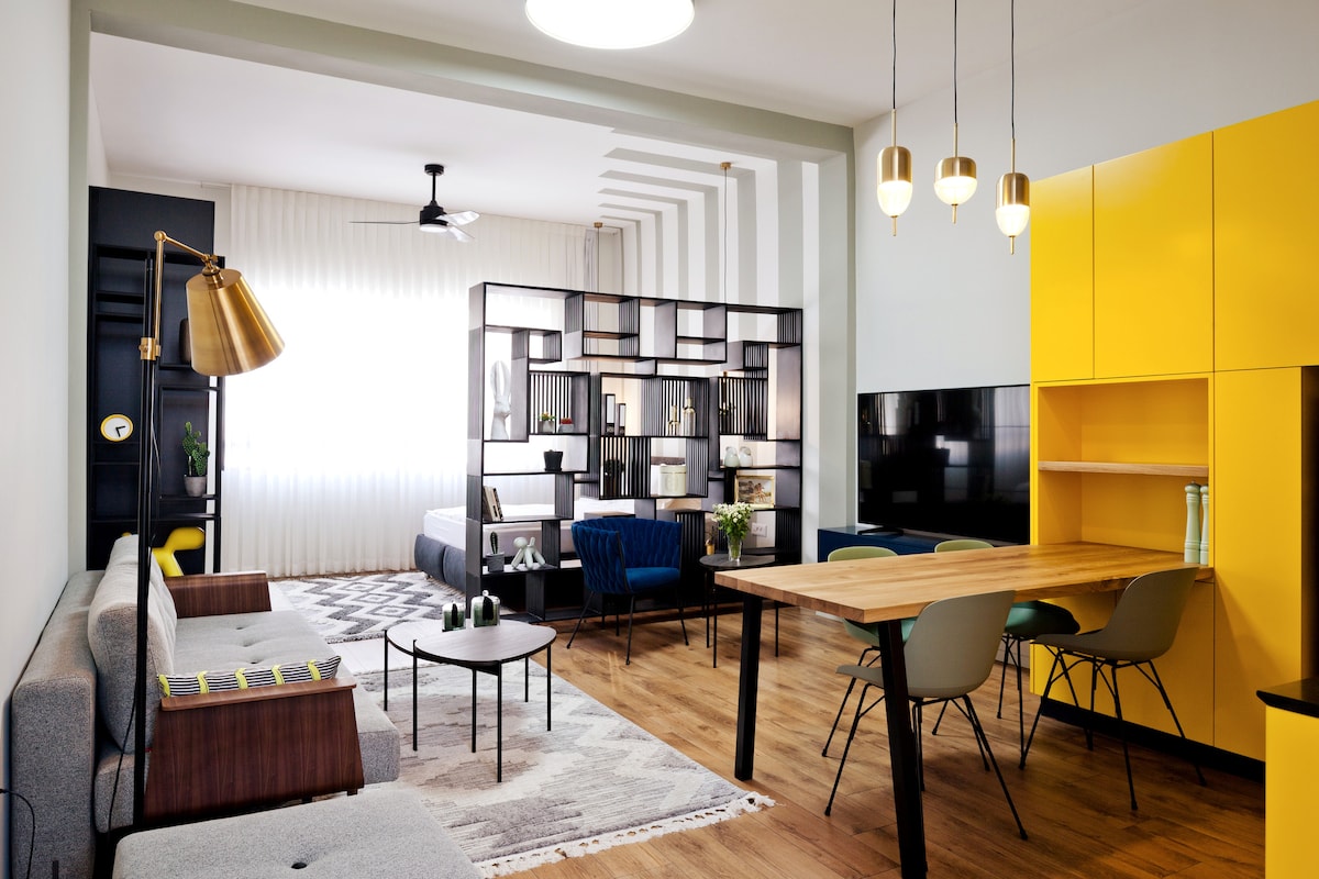 A stylish living area features a gray sofa and a custom-made sofa bed, separated by a modern black partition. A yellow cabinet stands beside a dining table surrounded by colorful chairs. A 65-inch flat-screen smart TV is wall-mounted, with natural light illuminating the space.
