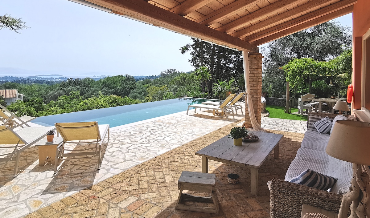 A spacious outdoor living area is featured, showcasing comfortable seating and a dining table under a wooden pergola. An infinity pool stretches beyond, reflecting the greenery surrounding the property. Sun loungers are positioned near the pool, providing a serene view of the landscape.
