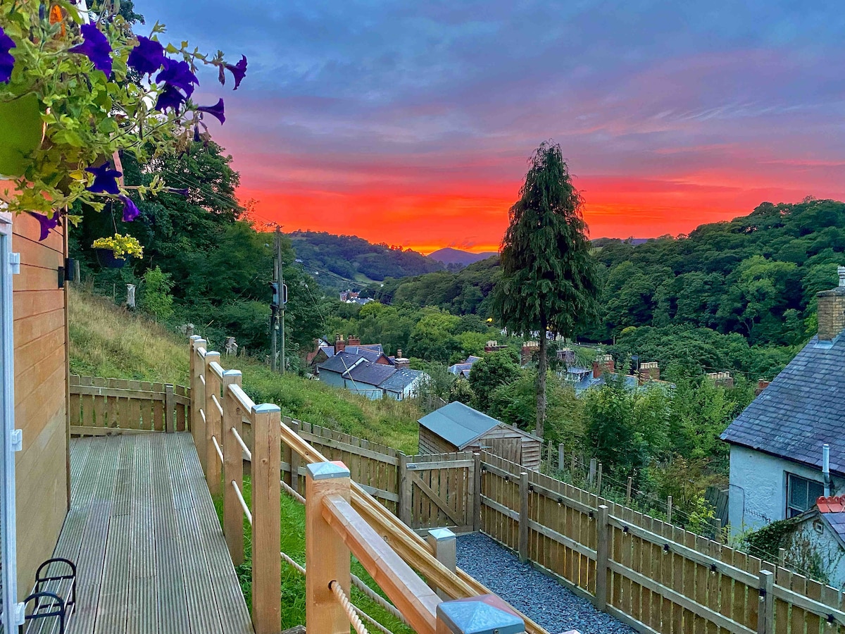 A wooden deck offers a scenic view of the vibrant sunset over the lush valley, framed by trees. Flowering plants hang from the railing, enhancing the natural beauty of the outdoor space. The surrounding hills and houses are illuminated by the warm evening light.