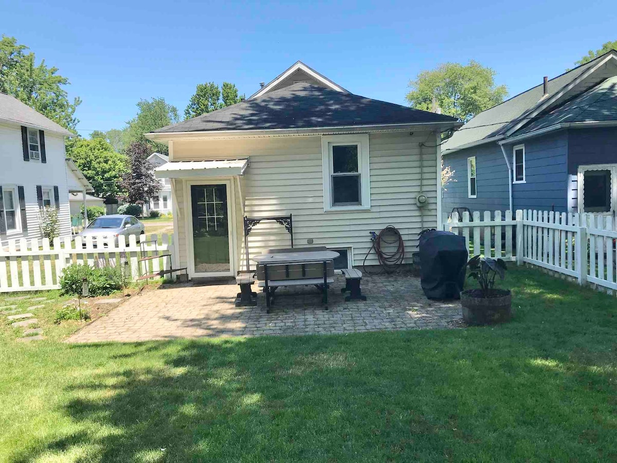 A fenced backyard features a stone patio with a picnic table and a grill. Lush green grass surrounds the area, complemented by a small garden space. The home's back entrance is visible, showcasing a door framed by two windows, with white fencing enclosing the yard.