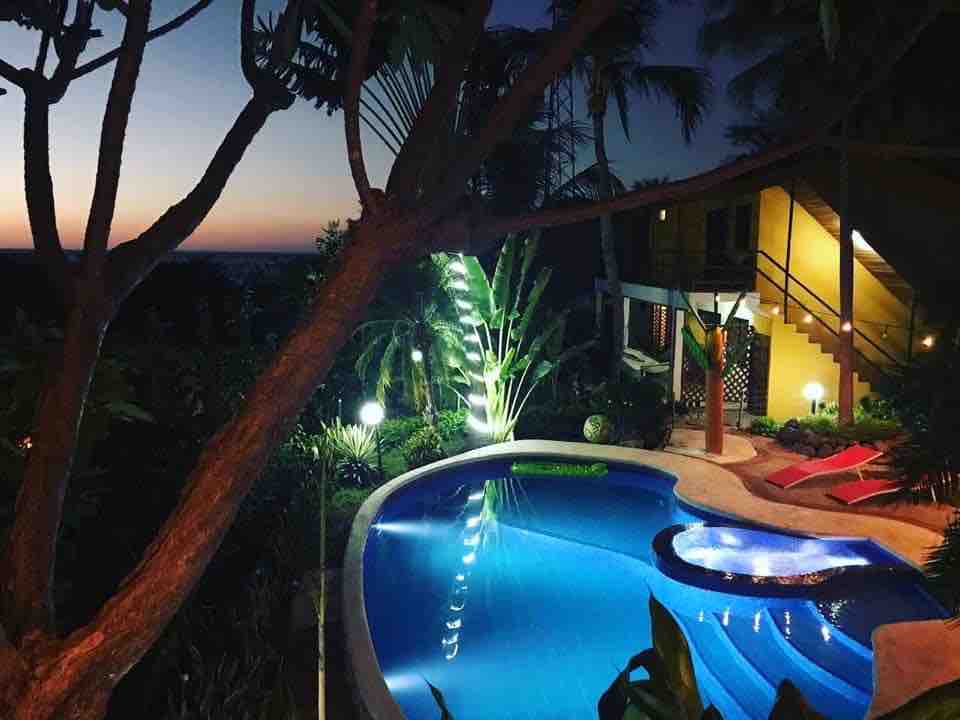 A tranquil pool area is illuminated by soft lighting, with a jacuzzi integrated into the design. Lush vegetation surrounds the pool, adding natural beauty. The sky transitions from vibrant colors to twilight, highlighting the serene atmosphere of the setting.