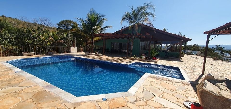 A private outdoor area features a large pool with crystal-clear water, surrounded by tropical plants and stone pathways. The spacious deck is complemented by a covered seating area, offering a relaxing environment against a backdrop of green hills.