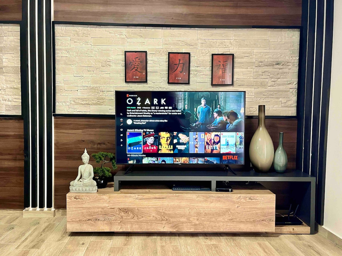 A modern living room is depicted with a 55-inch smart TV displaying a show from Netflix. Below the TV, a wooden console holds decorative vases and a small statue. The wall features a decorative design with framed artwork, complemented by wood paneling.