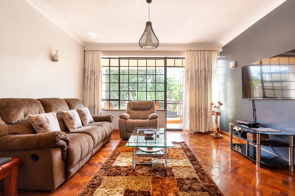 A cozy living area is highlighted by soft brown recliner seats and a plush carpet. Large windows allow natural light to fill the space, while a flat-screen TV is mounted on the wall. A glass coffee table complements the inviting furnishings.