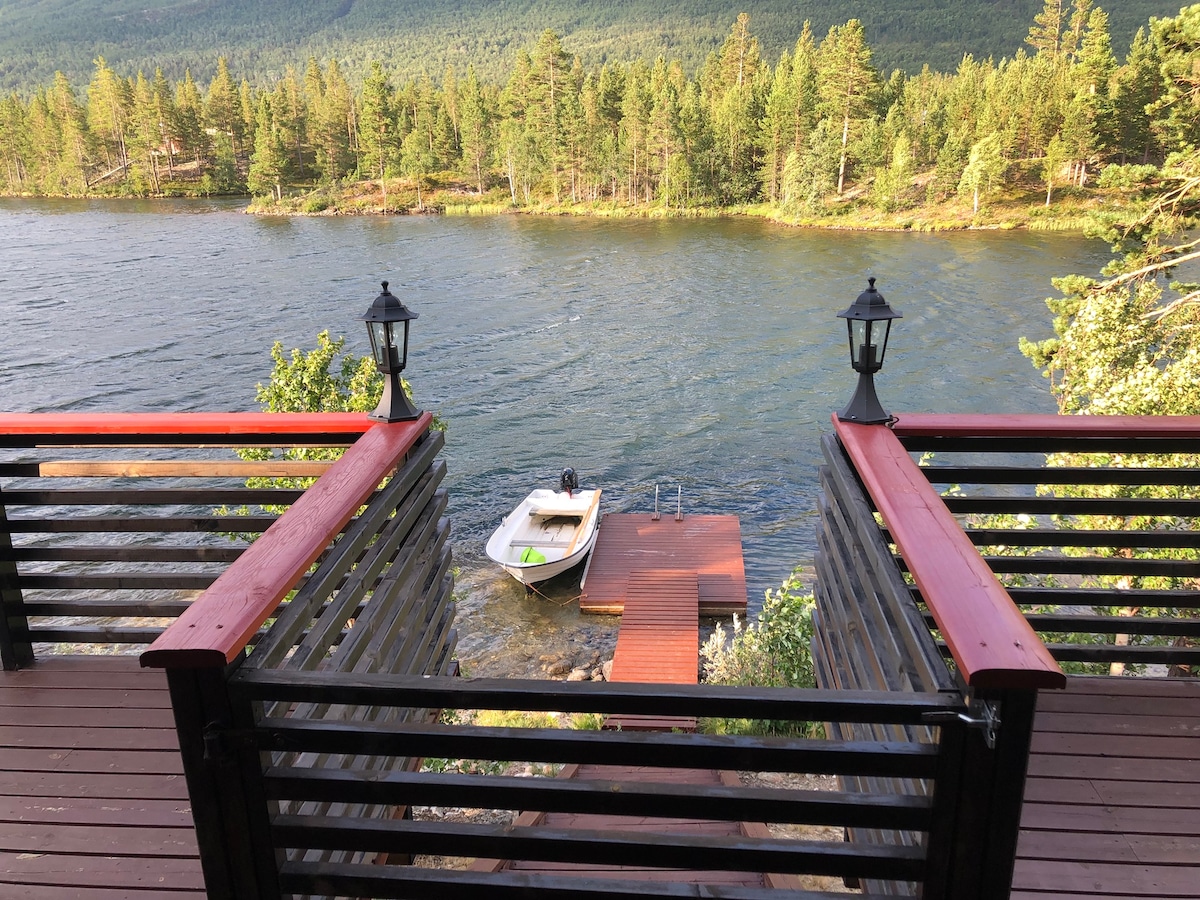 A wooden terrace features a view overlooking the calm waters of Lake Lesjaskogsvatnet. A small boat is moored at a wooden dock, complemented by two lanterns on either side of the staircase leading down to the water.