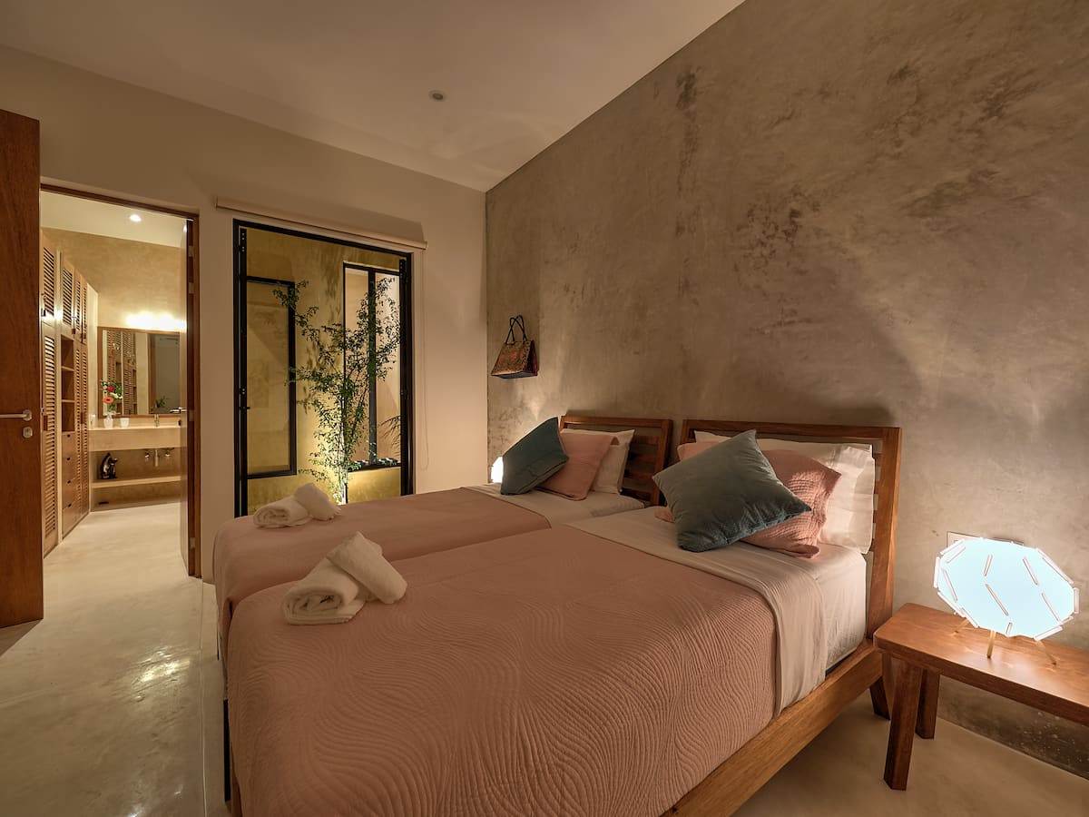 Chambre à coucher moderne avec deux lits simples, décorée avec des coussins à motifs et une lampe de chevet ajoutant une touche contemporaine, vue sur une petite cour intérieure.