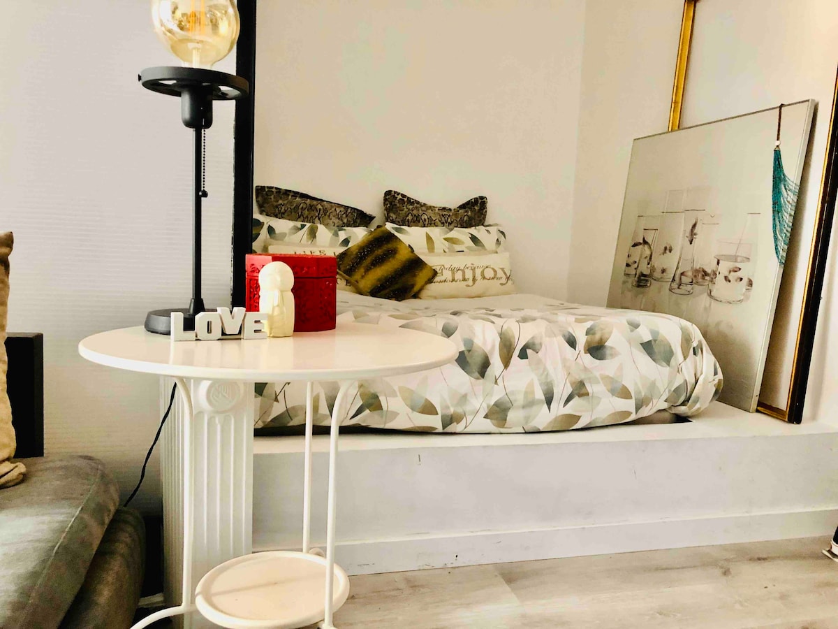 A cozy sleeping area is highlighted by a spacious bed adorned with decorative pillows and a patterned duvet cover. A small round table, accompanied by a lamp, is positioned nearby. A decorative piece with a calming aesthetic is seen in the background.