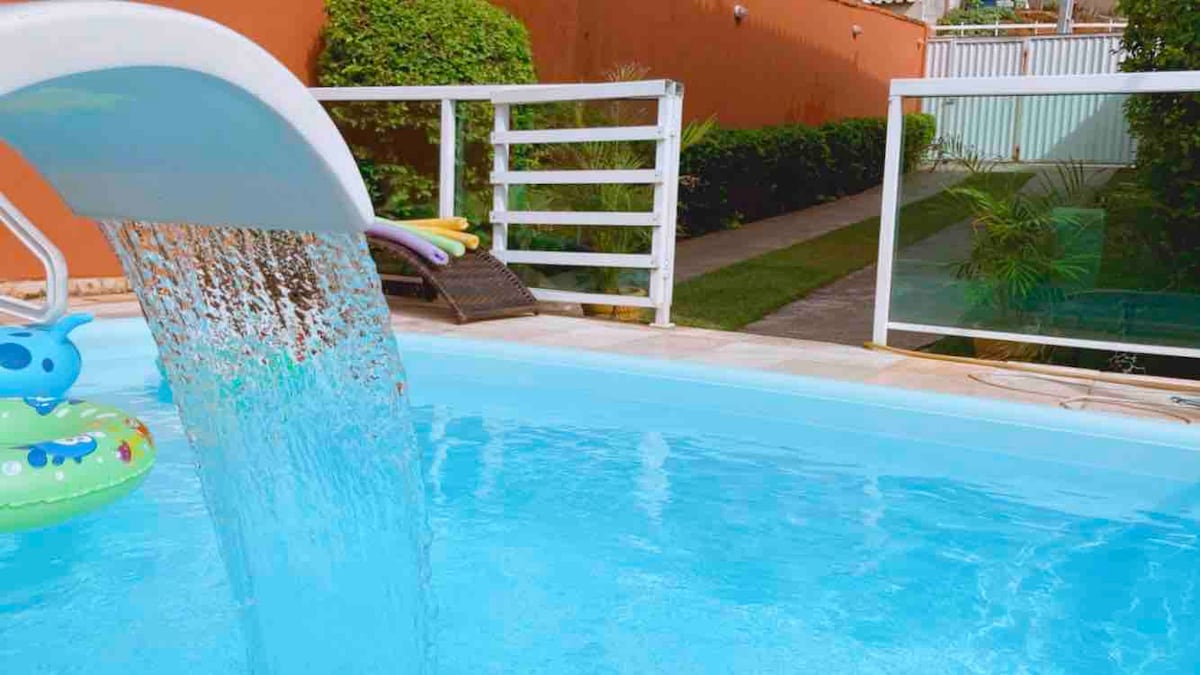 A private pool is featured, with clear blue water cascading from a slide. Lush greenery surrounds the area, offering a serene backdrop, while a pathway leads to additional outdoor space. Colorful inflatables are visible beside the pool, enhancing the inviting atmosphere.