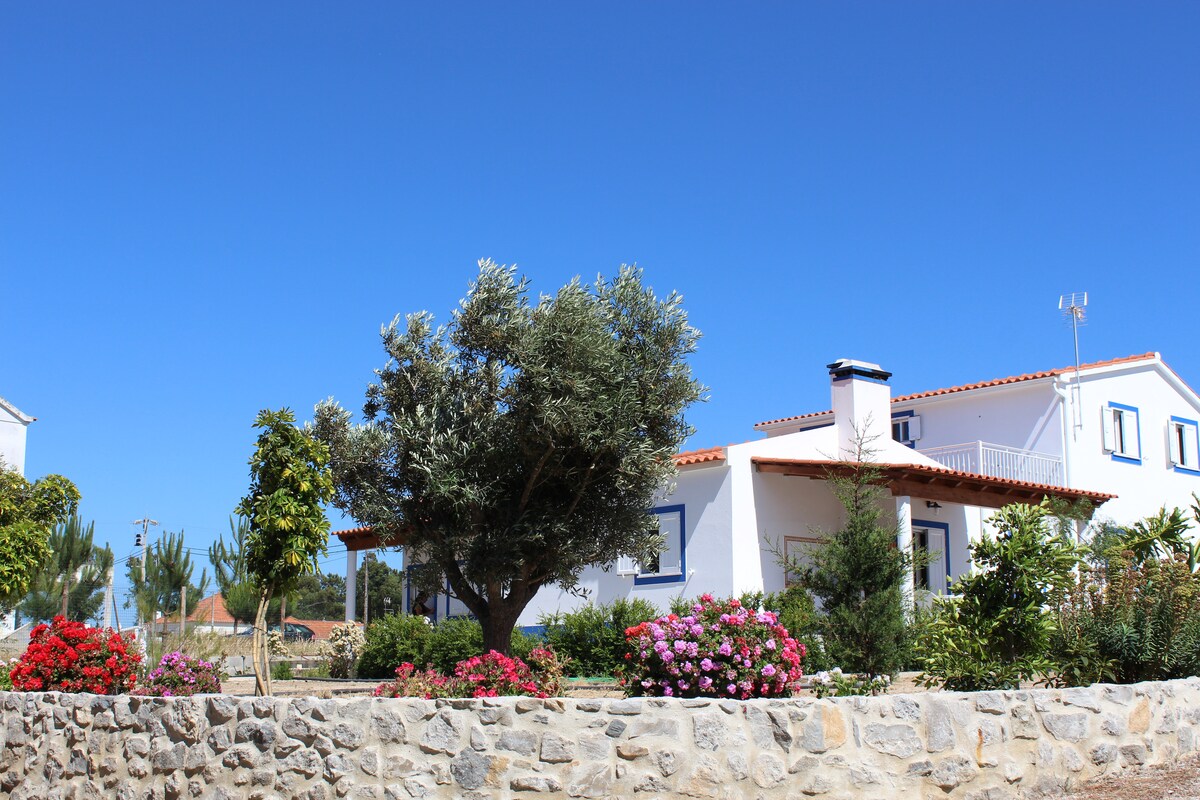 A two-story house is surrounded by vibrant gardens featuring flowering shrubs and trees. A stone wall encloses the property, while a clear blue sky provides a bright backdrop. The building showcases a traditional design with a red-tiled roof and multiple windows.