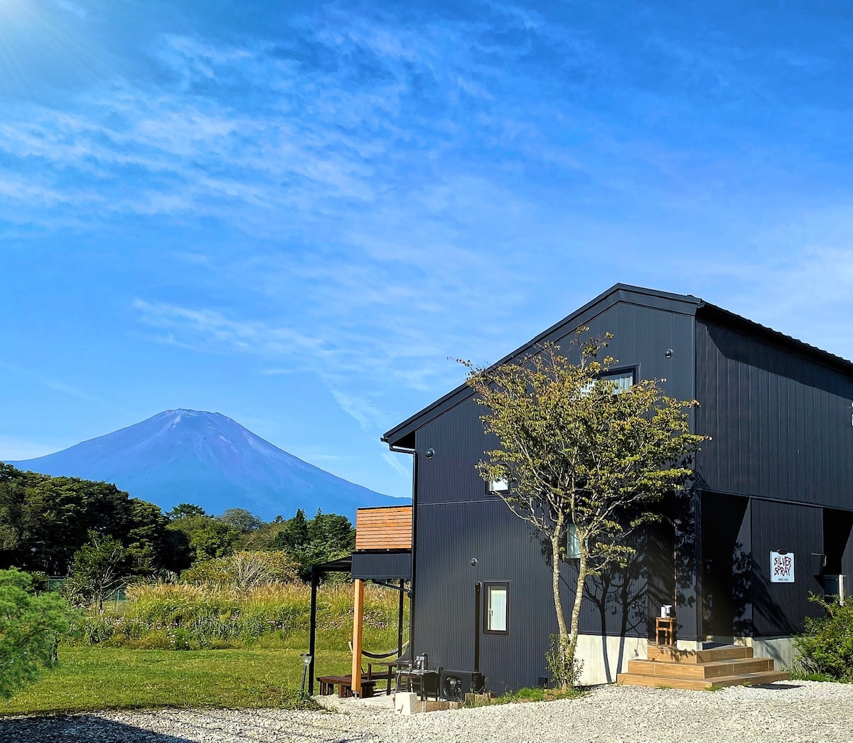 A modern, dark-colored cottage is situated against a backdrop of Mount Fuji. The structure features large windows and a wooden deck. Lush greenery surrounds the area, with a gravel pathway leading up to the entrance.