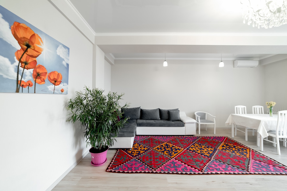 A bright living area is softened by a vibrant area rug featuring intricate patterns. A sectional sofa is placed against the wall, complemented by a white dining table set for six. A floral painting decorates one wall, while natural light fills the space.