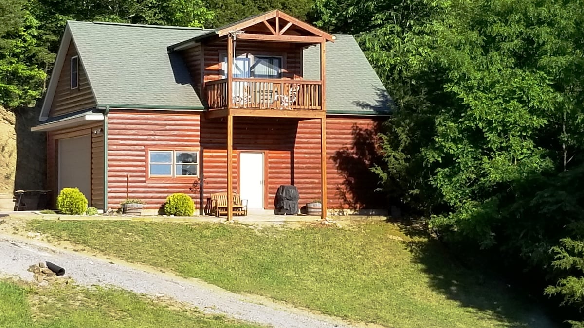 A rustic cabin is set on a gentle slope, featuring a wooden façade and a pitched roof. A small porch is accessible from the second floor, framed by a triangular overhang. Landscaping includes bushes and gravel paths leading to the entrance.