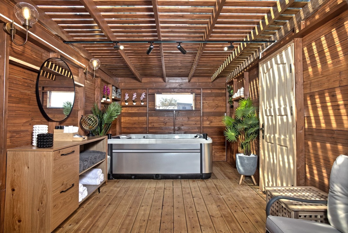 A modern spa area features a sleek jacuzzi with a wooden deck surrounded by natural elements. Decorative shelving displays rolled towels and plants, while soft lighting enhances the wood-paneled ambiance. A comfortable seating option is positioned nearby for relaxation.