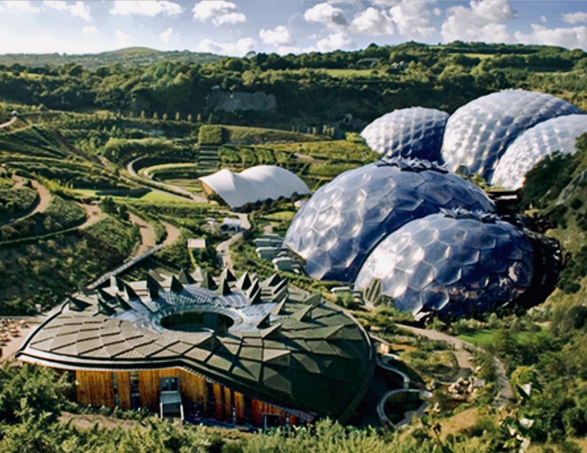 A striking aerial view of the Eden Project displays multiple geodesic dome structures housing diverse plant species. Lush greenery surrounds the domes, with winding pathways and landscaped gardens integrating into the natural hillside. The structure's distinct architectural features blend harmoniously with the unique landscape.
