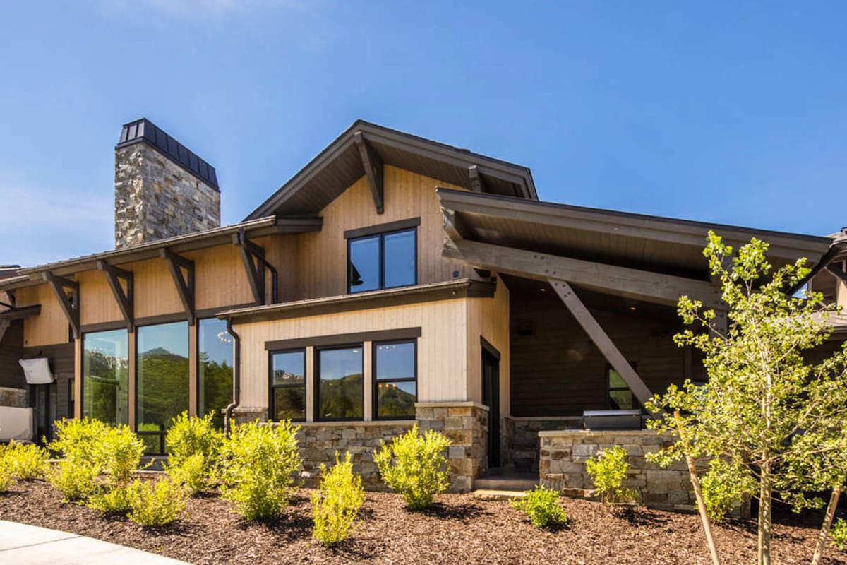 The exterior of a spacious home features a combination of wood and stone elements. Large windows allow natural light to fill the interior, while a chimney adds character. Surrounding the house, carefully landscaped greenery enhances the inviting entrance.