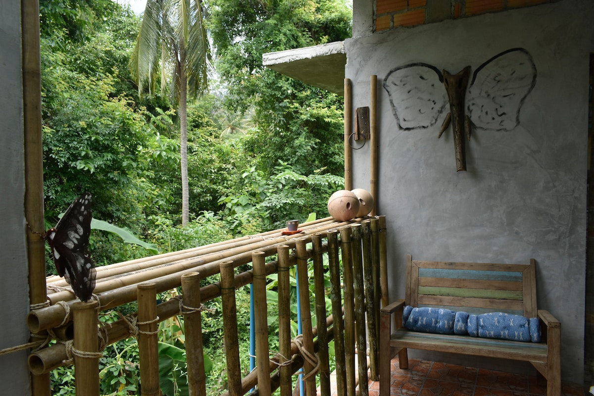 A natural balcony is bordered by bamboo railings, offering views of lush greenery. A wooden bench with blue cushions provides a cozy seating option. Two butterfly motifs are mounted on the rustic wall, enhancing the connection to nature.