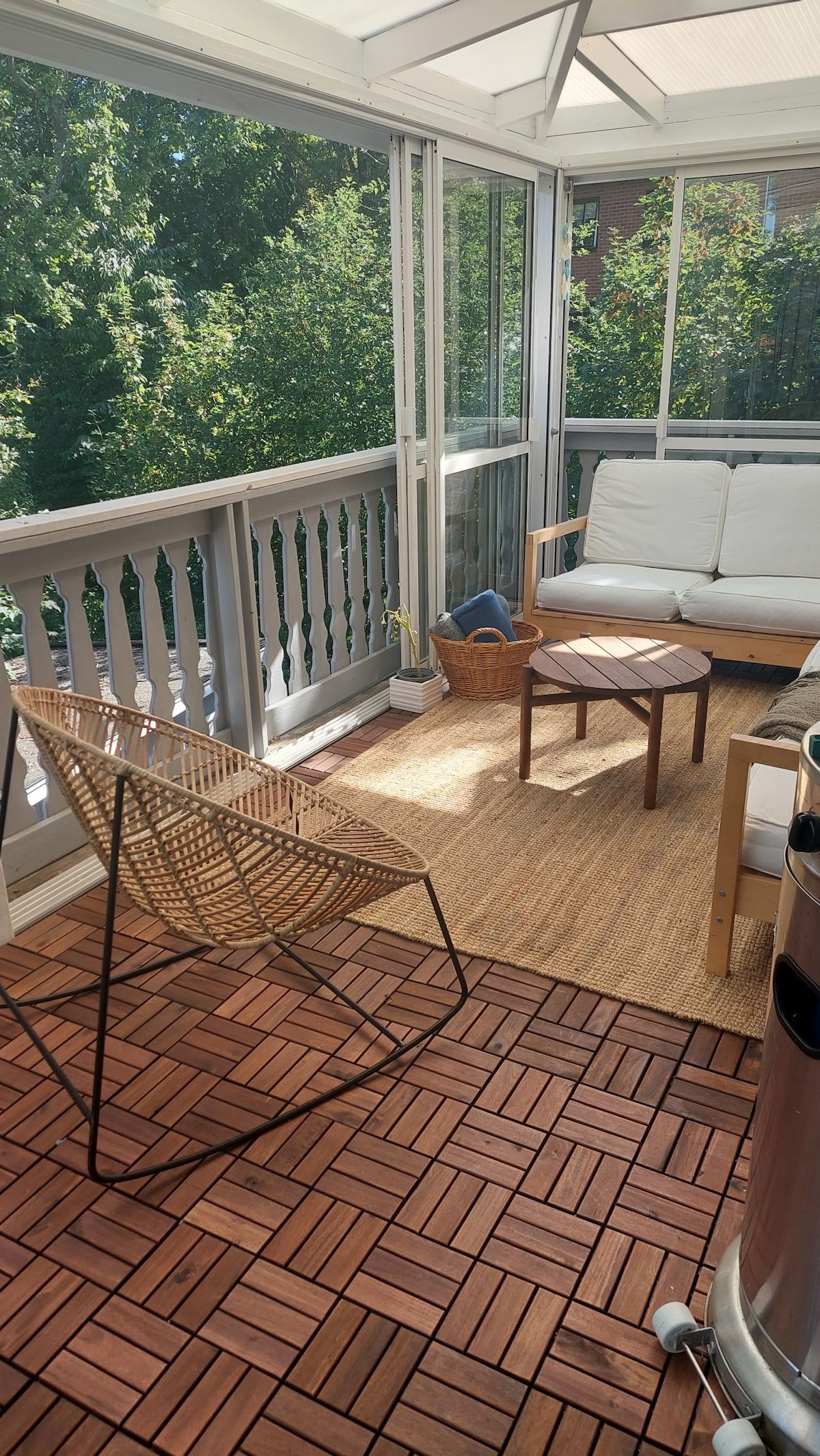 A sunlit balcony features a comfortable seating area with a light-colored sofa and a small coffee table. A woven chair adds texture against the wooden flooring. Lush greenery is visible through the glass walls, creating a refreshing outdoor-indoor connection.