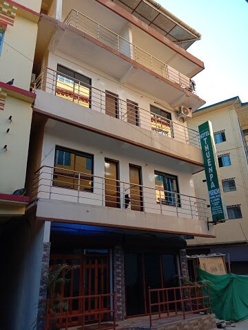 The exterior of a three-story hotel building features a light-colored facade with multiple large windows. A balcony runs along the second floor, and the entrance is framed by stone accents. A green hotel sign is displayed prominently next to the entrance.