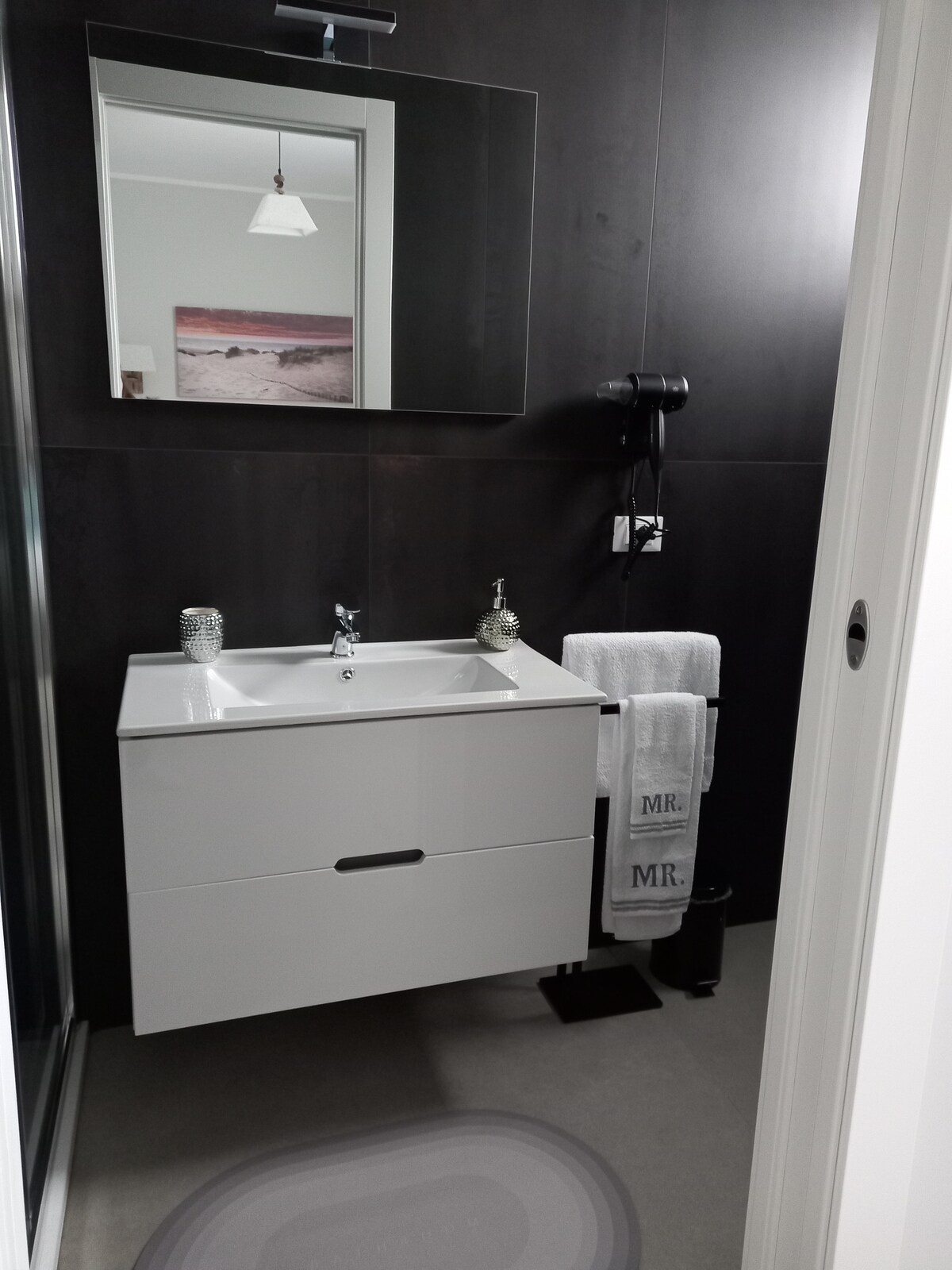 A modern bathroom features a white wall-mounted sink with a sleek cabinet below. Complimentary toiletries are neatly arranged on the countertop. A large mirror reflects the space, while a hairdryer is mounted nearby. Soft towels hang beside a stylish black waste bin.