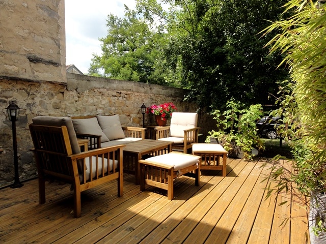 A wooden terrace is shown, furnished with a seating area comprising two armchairs, a sofa, and a coffee table, all featuring light-colored cushions. Lush greenery and flowering plants surround the area, creating a private and serene atmosphere.