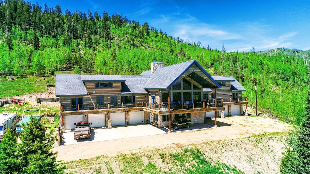 A large, well-constructed building is set against a backdrop of lush green trees under a clear blue sky. The structure features a spacious covered porch with wooden railings, providing ample outdoor space. Vehicles are parked in the driveway, which extends along the front of the property.