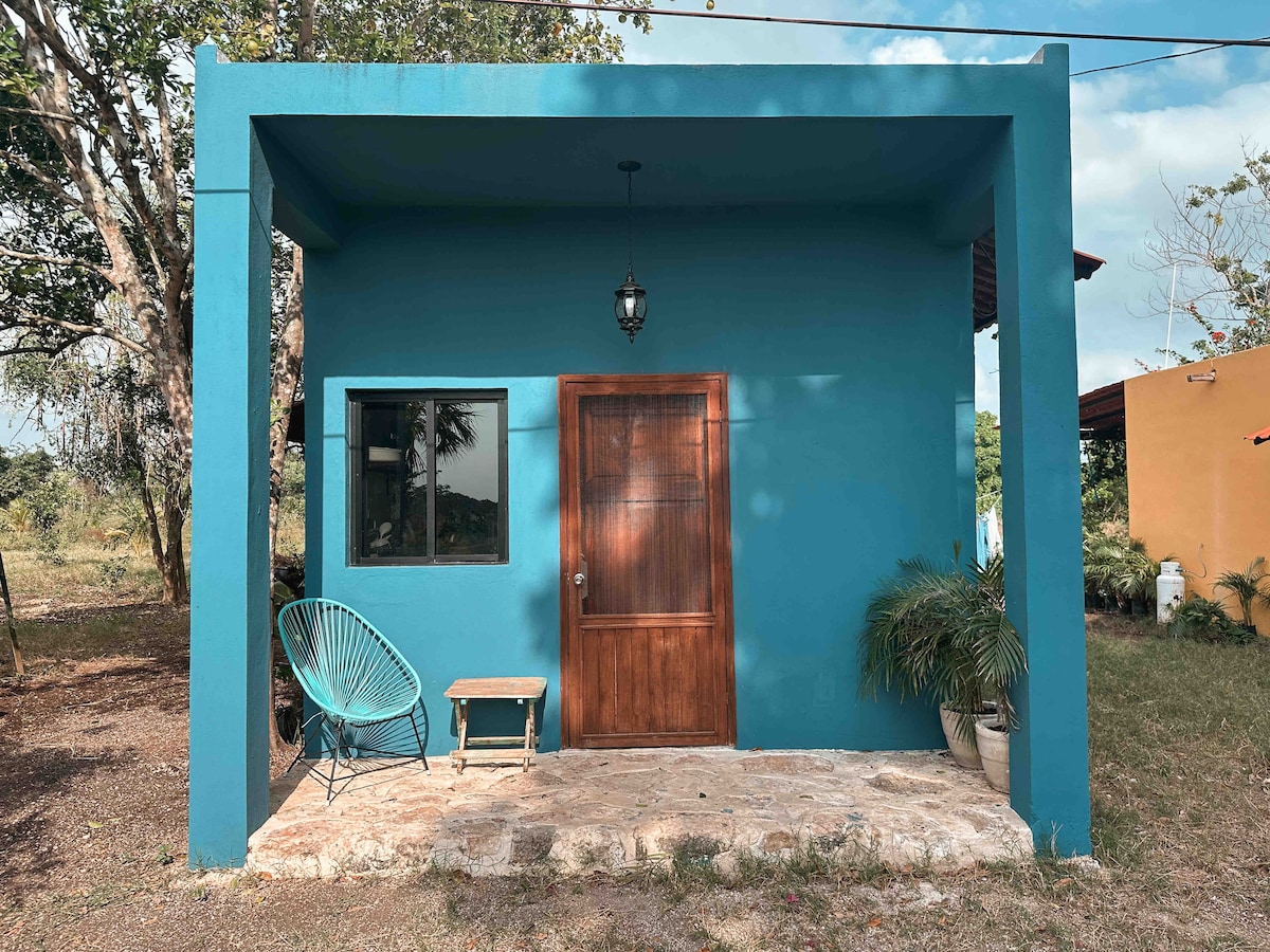 A vibrant blue exterior highlights the entrance of a cozy building, featuring a wooden door and a large window. A small wooden table and a green chair are positioned on the stone patio, surrounded by a natural landscape.
