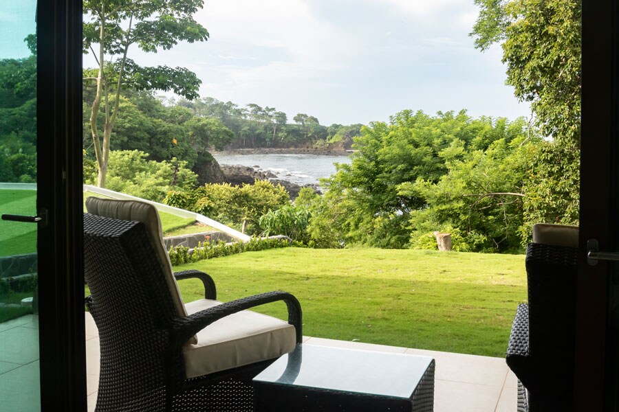 A serene outdoor space is depicted, featuring a comfortable chair with a light cushion alongside a small table. Lush greenery surrounds the area, leading to a view of the rocky shoreline and distant trees. Bright natural light filters into the scene, enhancing the tranquil environment.