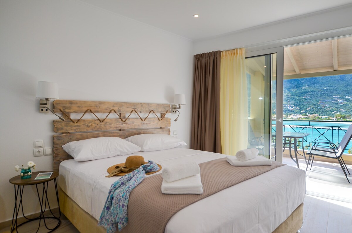 A bright and airy room features a spacious bed with white linens, complemented by a wooden headboard. A pair of sliding glass doors leads to a terrace with views of the gulf. Subtle decor elements include a small table and neatly arranged towels.