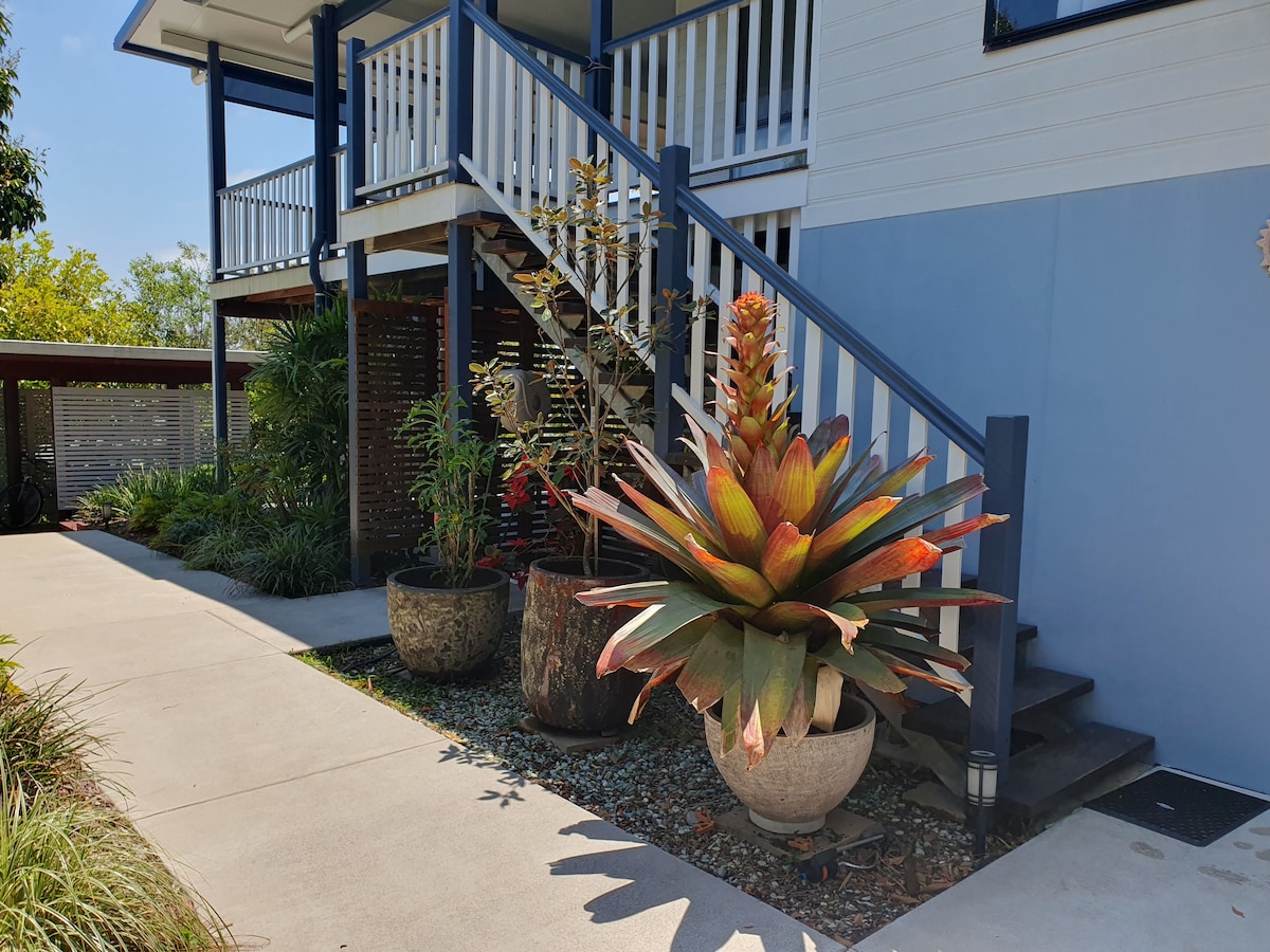 A well-maintained entrance area is highlighted by a large, colorful plant in a decorative pot. The pathway leads up to a set of stairs, framed by lush gardening and potted greenery, enhancing the welcoming atmosphere of the property.