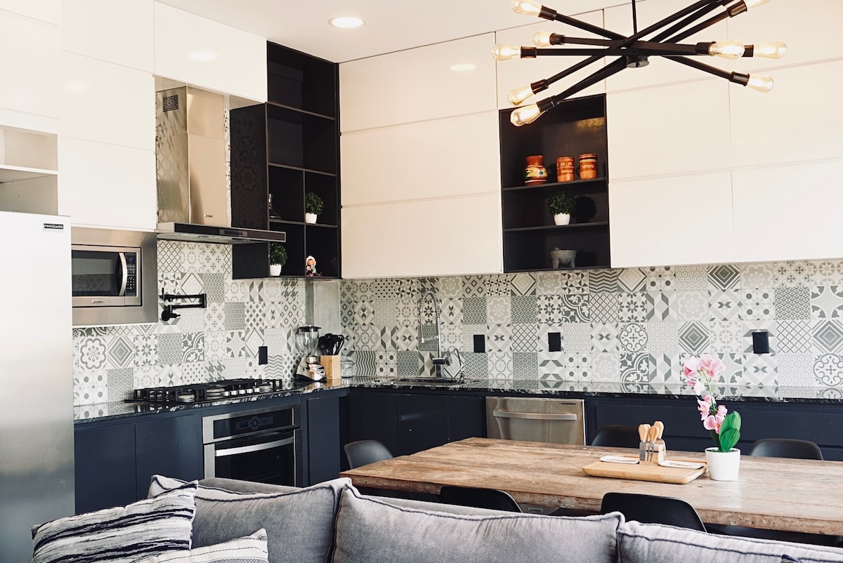 A modern kitchen features a blend of black and white cabinetry, accented by decorative tile backsplash. Stainless steel appliances and a gas stove are visible, complemented by open shelving displaying decorative items. A wooden dining table with seating is positioned near a plush sofa.