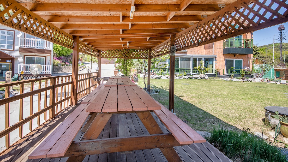A spacious outdoor seating area is featured under a wooden pergola, providing ample shade. Long wooden picnic tables are arranged, suitable for group gatherings. Lush green grass and elements of landscaping are visible in the surrounding area, creating a relaxed outdoor environment.