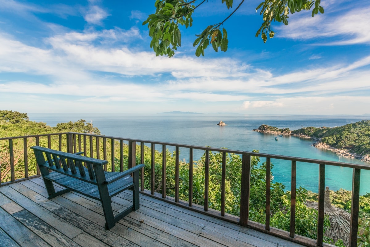 A wooden deck features a simple bench, overlooking clear turquoise waters and rocky coastlines. Lush greenery surrounds the area, with a bright blue sky and soft clouds completing the serene view. The peaceful setting invites relaxation and enjoyment of the natural beauty.
