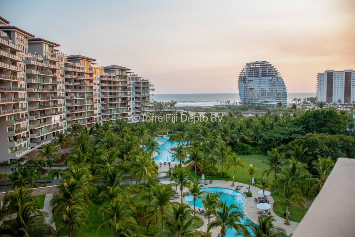An expansive view of a luxurious residential complex is visible, featuring multiple buildings and lush greenery. The beachfront can be seen in the background, with a serene sky transitioning into twilight colors. Shimmering pools are framed by palm trees in the foreground.