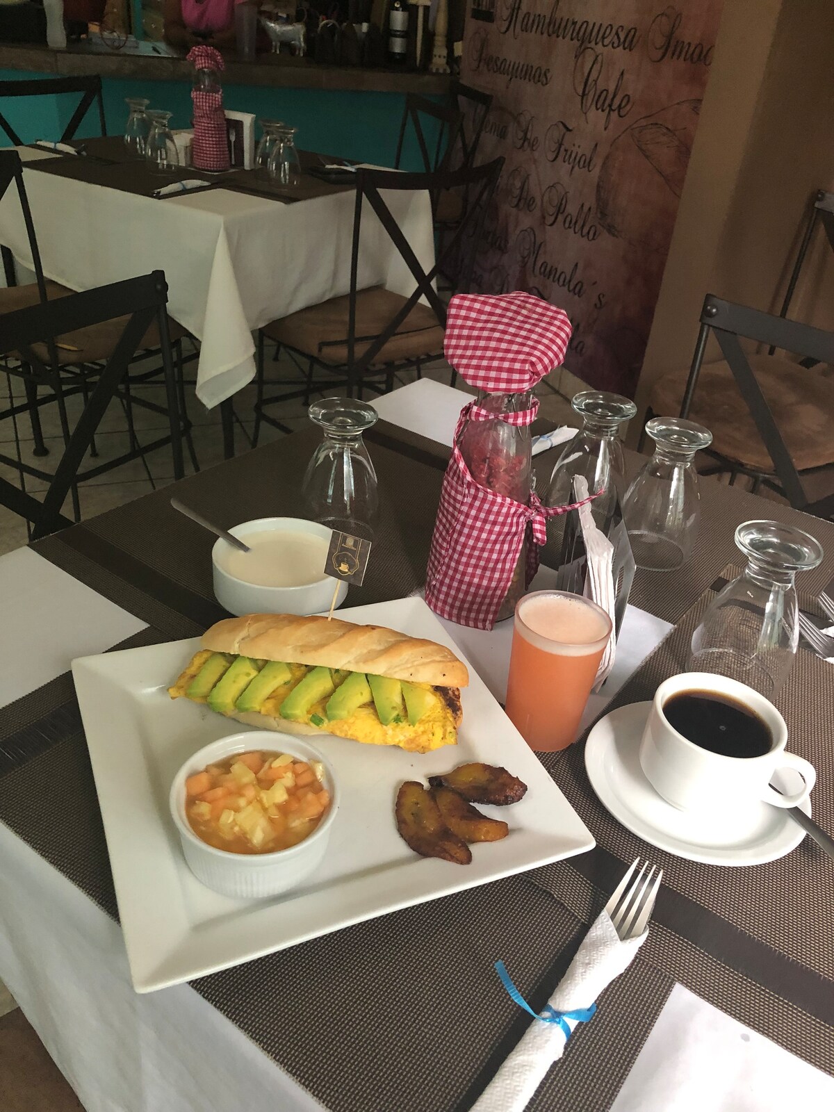 A breakfast plate is presented on a white square dish, featuring an omelette-filled sandwich with slices of avocado. Accompanying items include a side of diced fruit, crispy plantains, and a small bowl of sauce. A cup of coffee and a refreshing drink are placed on the table.