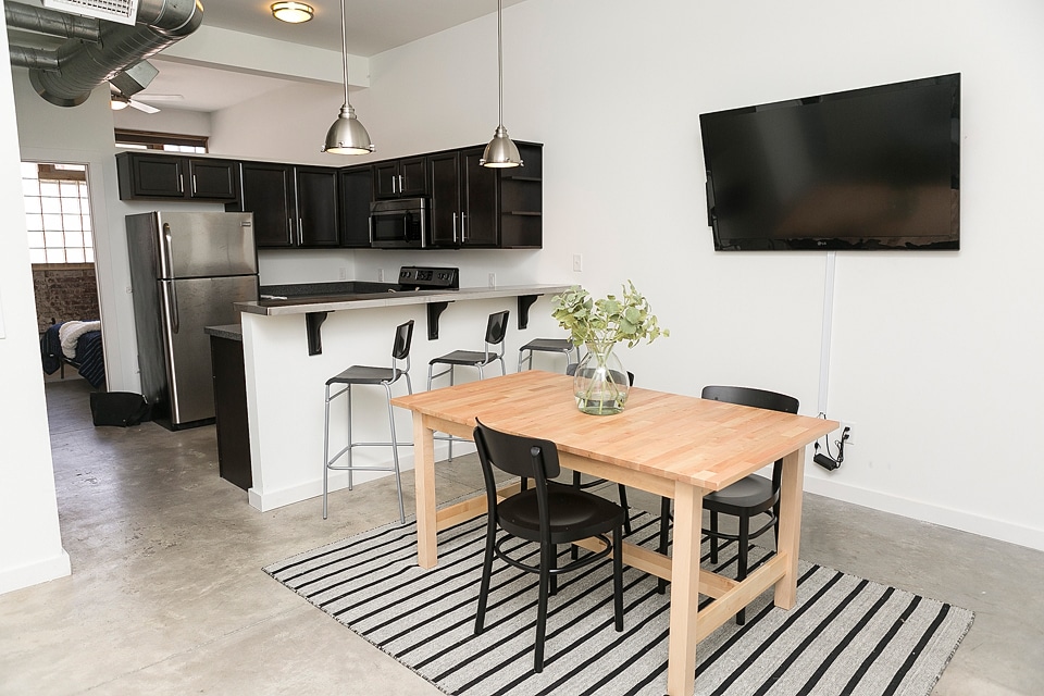 An open-concept dining area showcases a simple wooden table with four black chairs. A large area rug with stripes is laid beneath the table. The kitchen features dark cabinetry and stainless steel appliances, while a wall-mounted TV is positioned nearby for entertainment.