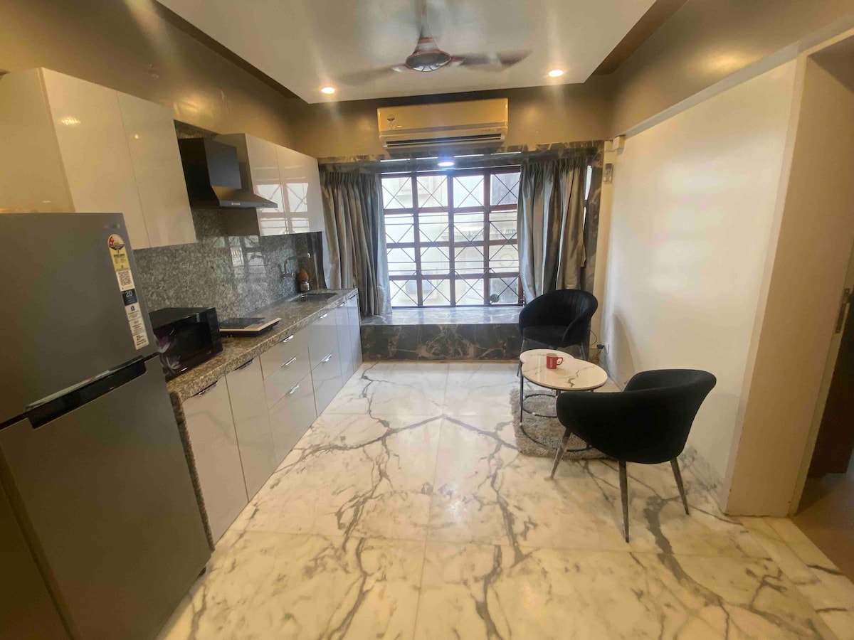 A modern kitchen area is depicted with sleek cabinetry and a granite countertop. The space is equipped with a refrigerator, microwave, and a small dining table with two black chairs. Natural light filters through a large window, brightening the marble flooring.