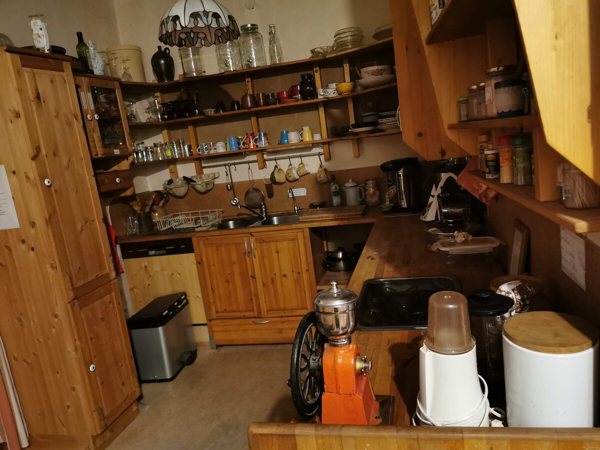 A warm kitchen environment showcases wooden cabinetry and shelves lined with dishes and utensils. A mix of appliances is visible, including a coffee grinder and a stand mixer. Counterspace provides areas for food preparation, while a window allows natural light into the room.