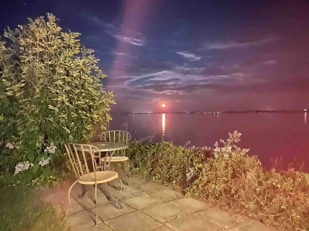 A small patio area features a round table with two chairs, set against a backdrop of a serene river. The glowing reflection of the moonlight dances on the water's surface, while nearby greenery frames the view, creating a peaceful outdoor setting.
