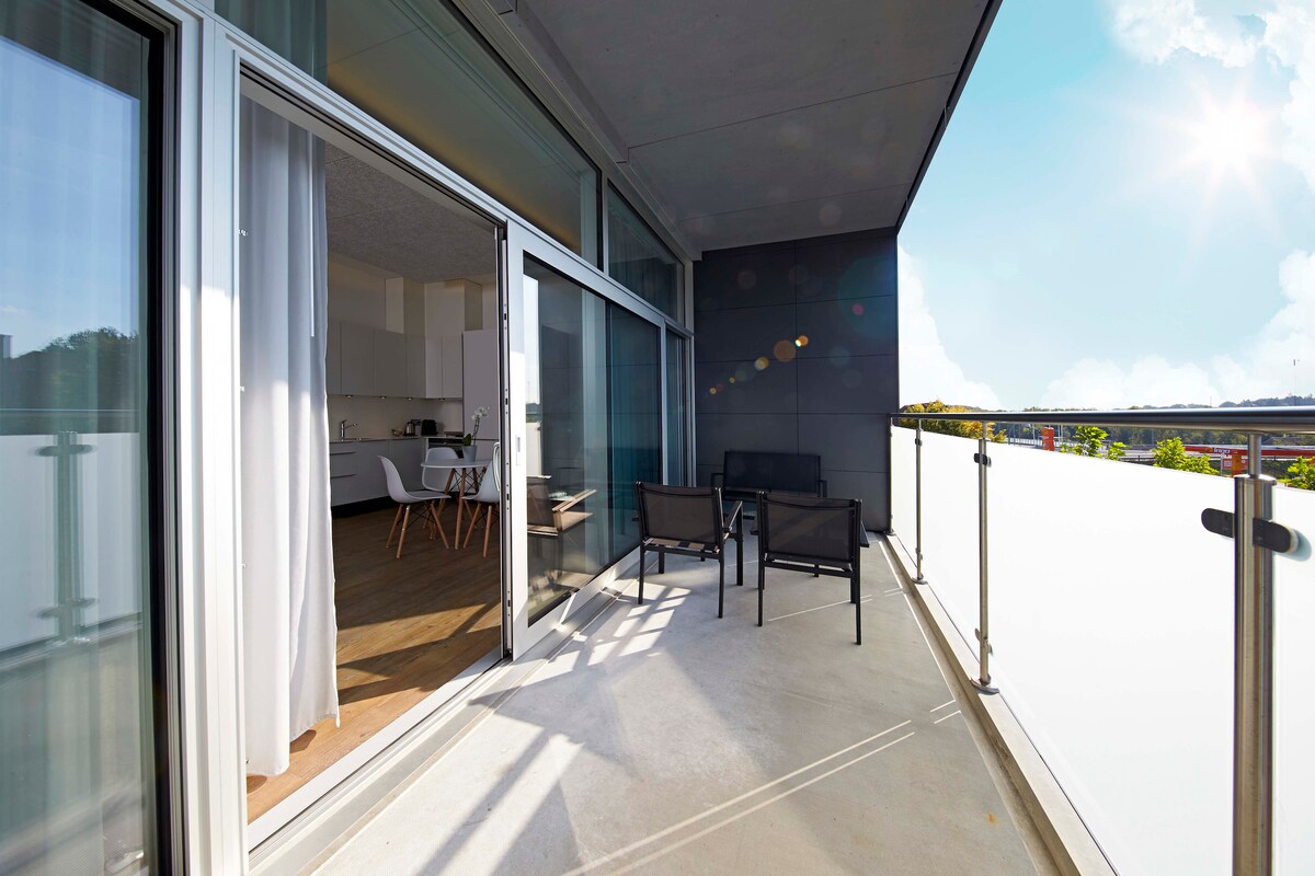 An expansive balcony is showcased, featuring modern outdoor seating. Glass doors open to reveal a bright interior, allowing natural light to filter through. The balcony offers a view of the surroundings, complemented by a clear blue sky.