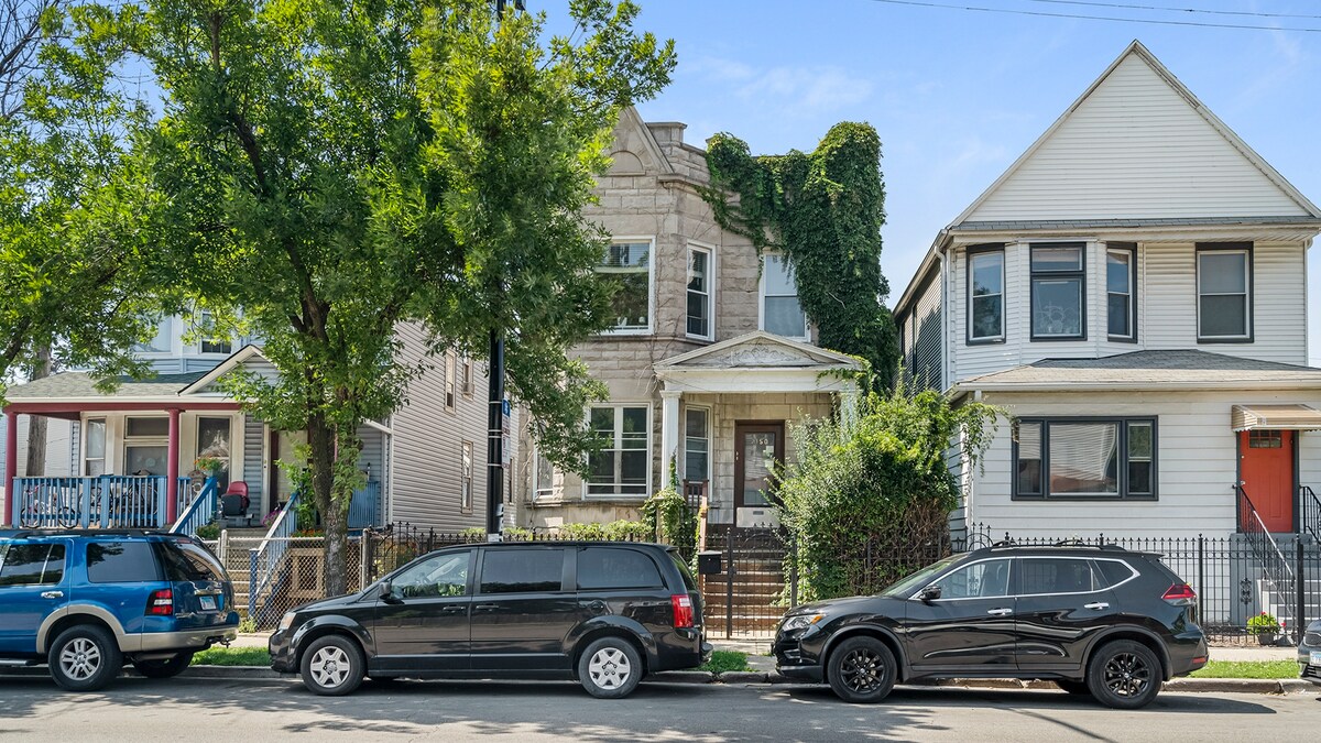 A two-story greystone building is positioned amidst trees and neighboring homes. The entrance features a porch with a few steps leading to the front door, which is framed by greenery. Vehicles are parked along the street, showcasing a typical urban setting.