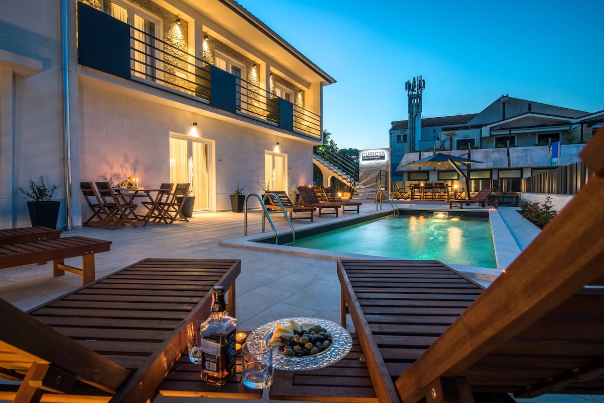 The outdoor area features a pool surrounded by lounge chairs, creating a relaxing space for leisure. Soft lighting illuminates the modern building and the patio, while tables and chairs are arranged for outdoor dining. Greenery is present, enhancing the tranquil ambiance.