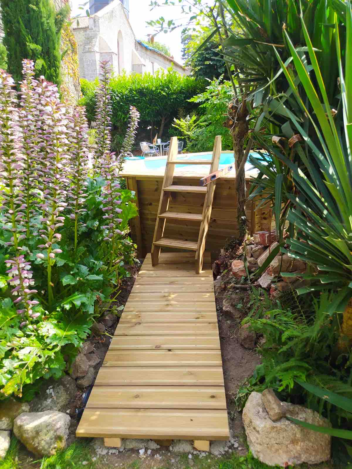 A wooden pathway leads to a pool, surrounded by lush greenery and vibrant flowering plants. A wooden ladder is positioned at the edge of the pool, providing access. The natural setting is enhanced by tall foliage and a serene ambiance.