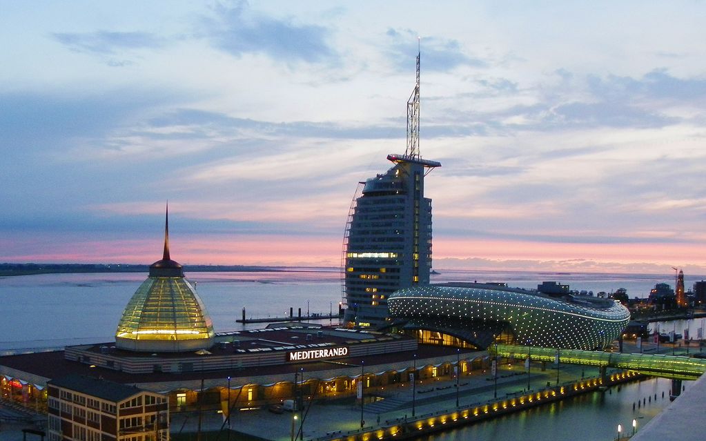 A scenic view of the Mediterraneo building and surrounding structures is captured during dusk. The sky shows soft pastel hues, reflecting on the water's surface. The unique architecture features a dome and curved elements, blending with the tranquil atmosphere of the waterfront.