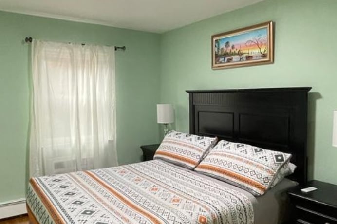 The bedroom features a spacious bed dressed in a patterned blanket. Light green walls complement the room, while natural light filters through sheer curtains. A framed artwork hangs above the bed, and bedside lamps offer additional illumination.