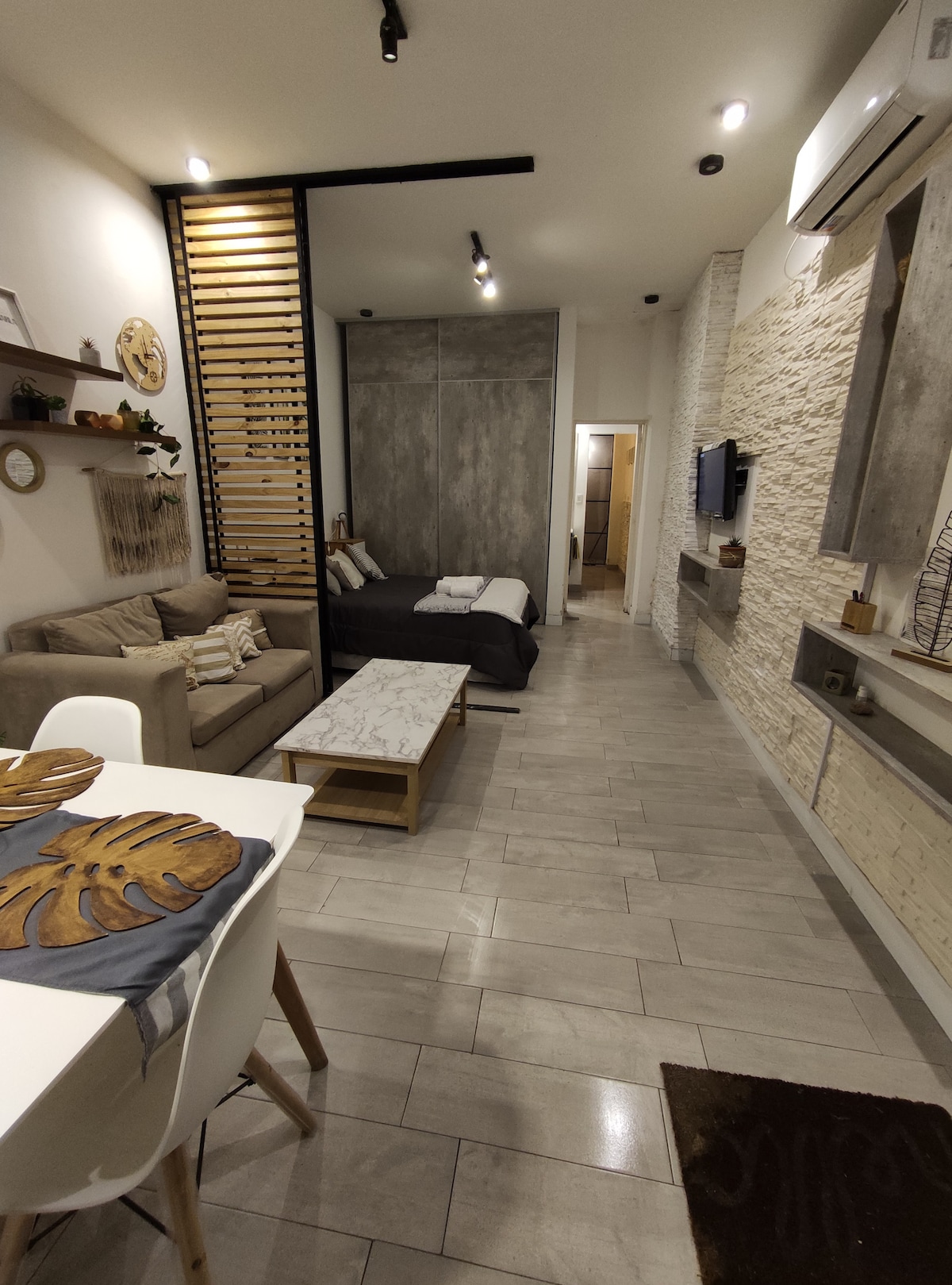 A functional living space is divided by wooden slats, with a modern sofa and spacious bed visible. The dining area features a light wood table and white chairs, while textured walls and ambient lighting create a warm environment. A TV and air conditioning unit are present.