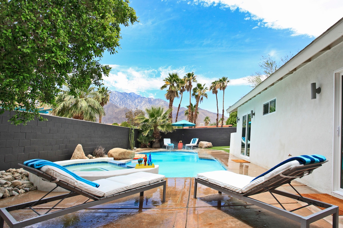 A serene outdoor space features a pool surrounded by palm trees and mountain views. Lounge chairs are positioned near the pool, while colorful floats and a small table add to the inviting atmosphere. A spacious patio area is designed for relaxation and enjoyment.