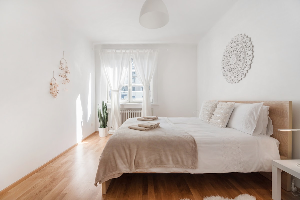 A bright bedroom features a spacious bed dressed in soft linens, with decorative cushions placed neatly on top. Natural light pours in through sheer curtains, illuminating the wooden floor. A potted plant adds a touch of greenery, enhancing the room's serene atmosphere.
