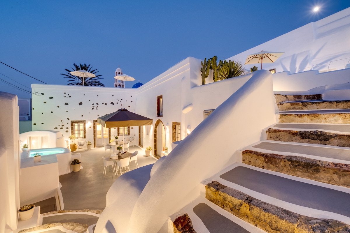 A whitewashed exterior is illuminated by soft lighting in the evening. Stone stairs lead to a multi-level space with an outdoor dining area featuring a parasol and decorative plants. A heated jacuzzi is visible, integrating nature and design harmoniously.