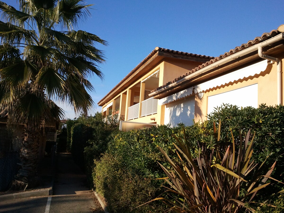 The exterior of a two-story residence is visible, featuring a peach-colored facade with a tiled roof. Lush greenery surrounds the building, including palm trees and various shrubs. Clear blue skies provide a bright backdrop, contributing to a serene outdoor environment.